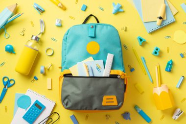 Okul malzemeleri konsepti. Üzerinde cetvel not defterleri olan açık sırt çantasının en üstteki fotoğrafı. Kalemler, dağınık kırtasiyeler ve izole edilmiş sarı arka planda içki şişesi.