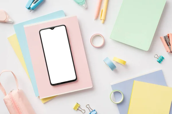 Back to school concept. Top view photo of smartphone over colorful diaries pens pencil-case staplers adhesive tape round correction tape and binder clips on isolated white background with blank space