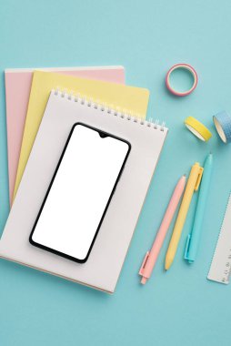 Back to school concept. Top view vertical photo of school supplies smartphone over stack of notepads pens ruler and adhesive tape on isolated pastel blue background with empty space