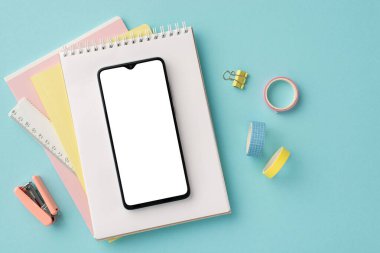 Back to school concept. Top view photo of school supplies smartphone over note pads ruler adhesive tape binder clip and mini stapler on isolated pastel blue background with empty space