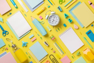 Alarm saati fotoğrafının üst görüntüsü. Renkli kırtasiye defterleri kalem zımbası, raptiye, hesap makinesi, cetvel alfabesi harfleri, yapıştırıcı makas keskinleştirici, sarı arkaplan.