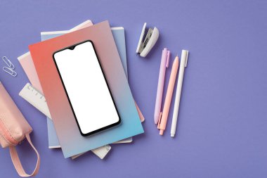 Back to school concept. Top view photo of smartphone over colorful copybooks pens stapler ruler clips and pink pencil-case on isolated purple background with empty space