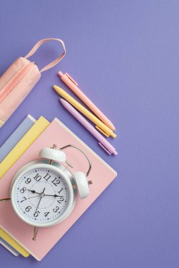 Okul konseptine geri dönelim. Renkli okul malzemelerinin en üstteki dikey fotoğrafı not defterlerinin üzerindeki alarm saati kalemler ve izole edilmiş menekşe arka plandaki pembe kalem kutusu.
