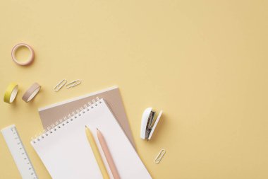 Okul aksesuarları konsepti. Kırtasiye fotokopi kitaplarının en üst görüntüsü cetvel kalemler zımba zımbaları ve izole edilmiş pastel sarı arkaplan üzerindeki yapışkan bantlar