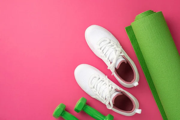 Sports concept. Top view photo of white sneakers green exercise mat and dumbbells on isolated pink background with empty space