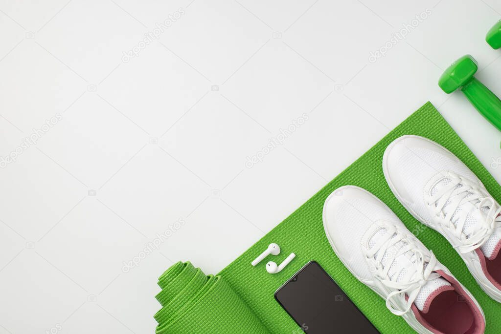 Fitness accessories concept. Top view photo of dumbbells white sneakers smartphone and earbuds over green exercise mat on isolated white background with empty space