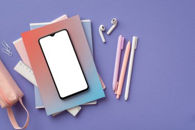Back to school concept. Top view photo of smartphone over colorful diaries earbuds pens ruler clips and pink pencil-case on isolated purple background with blank space