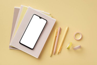 Back to school concept. Top view photo of smartphone over stack of copybooks adhesive tape and pens on isolated pastel yellow background with copyspace