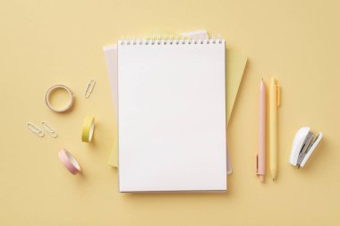 Okul konseptine geri dönelim. Okul aksesuarlarının en üst görünümü. Not defterleri, raptiyeler, zımba zımbaları ve izole edilmiş pastel sarı arka planda boş alan olan yapışkan bantlar.