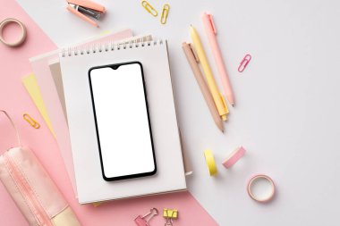 Back to school concept. Top view photo of school supplies smartphone over notebooks pens stapler binder clips adhesive tape and pencil-case on bicolor pink and white background with blank space