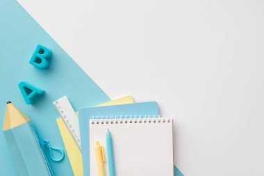 Okul konseptine geri dönelim. Sarı ve mavi okulun en üstteki fotoğrafı plastik alfabe harfleri, not defterleri, cetvel kalemleri ve iki renkli beyaz ve mavi arkaplan üzerine mavi kalem çantası, boş alan.