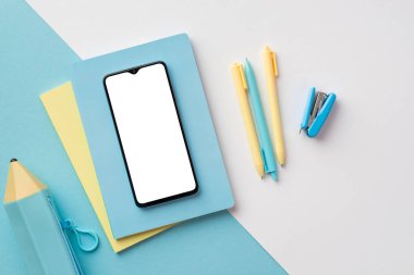 Back to school concept. Top view photo of smartphone over diaries pens stapler and blue pencil-case on bicolor blue and white background with copyspace