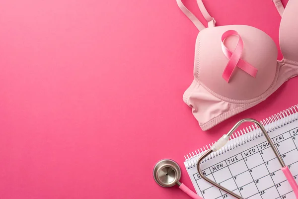 Breast cancer prevention concept. Top view photo of pink ribbon attached to brassiere calendar and stethoscope on isolated pink background with copyspace