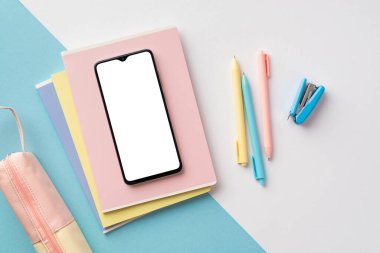 Back to school concept. Top view photo of school supplies smartphone over copybooks pens mini stapler and pink pencil-case on bicolor blue and white background with copyspace