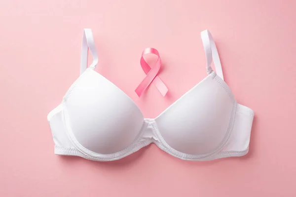 Top view photo of pink ribbon symbol of breast cancer awareness and white brassiere on isolated pastel pink background