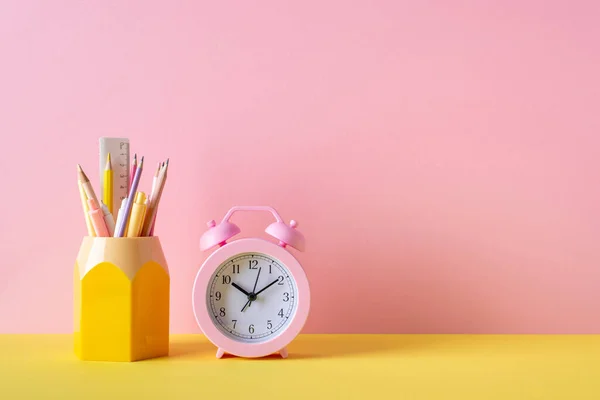 Back to school concept. Photo of school supplies on yellow desk pink alarm clock stand for pens pencils and ruler on pink wall background
