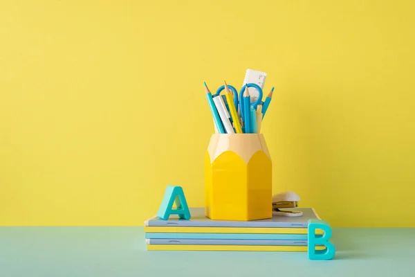 School accessories concept. Photo of stationery on blue desk plastic alphabet letters stack of notebooks stapler and stand for pens on yellow wall background