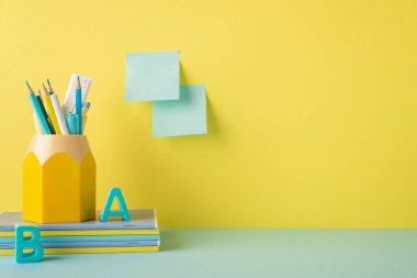 School accessories concept. Photo of stationery on blue desk stack of notebooks stand for pens plastic alphabet letters and sticky notes attached to yellow wall with copyspace