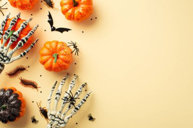 Halloween party decor concept. Top view photo of skeleton hands holding pumpkin creepy insects cockroaches centipedes spiders bat silhouette and confetti on isolated beige background with copyspace