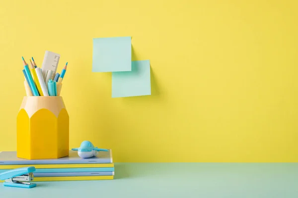 School accessories concept. Photo of stationery on blue table stack of notepads stand for pens stapler plane shaped sharpener and sticky notes attached to yellow wall with copyspace