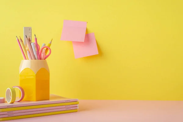 School supplies concept. Photo of trendy stationery on pink table stand for pens stack of copybooks adhesive tape and sticky note paper attached to yellow wall with copyspace