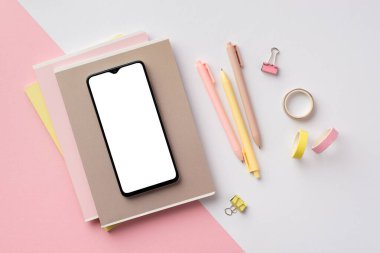 Back to school concept. Top view photo of smartphone over stack of copybooks pens binder clips and adhesive tape on bicolor pink and white background with empty space