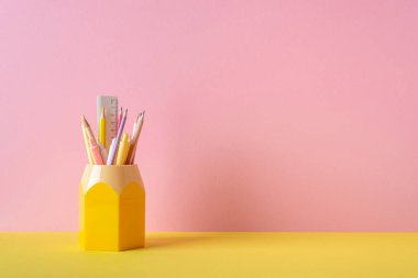 School supplies concept. Photo of stationery on yellow desk yellow stand for pens pencils and ruler on pink wall background with empty space