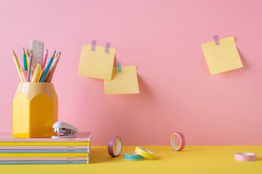 Back to school concept. Photo of school accessories on yellow table pencil holder stack of copybooks stapler adhesive tape and sticky note paper attached to pink wall