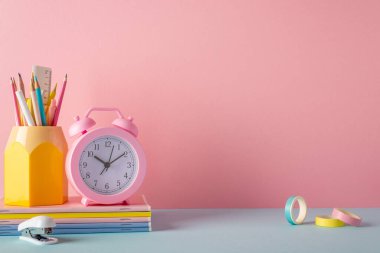 School accessories concept. Photo of stand for pencils alarm clock stack of notebooks mini stapler and adhesive tape on pink wall background