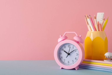 Back to school concept. Photo of school accessories on blue table pen holder alarm clock stack of notebooks and stapler on pastel pink wall background