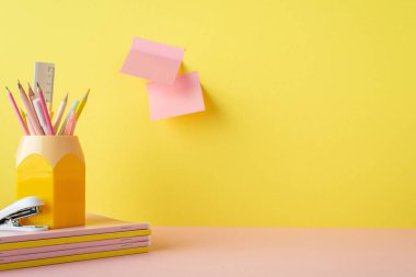 Back to school concept. Photo of pink and yellow school supplies stand for pencils stapler stack of copybooks and sticky note paper attached to yellow wall with copyspace