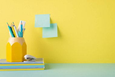 School accessories concept. Photo of stationery on blue tabletop stack of notebooks stapler stand for colour pencils and sticky notes attached to yellow wall