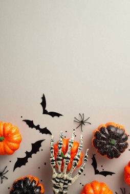 Halloween spooky decorations concept. Top view vertical photo of skeleton hand holding pumpkins bat silhouettes spiders and confetti on isolated grey background with copyspace