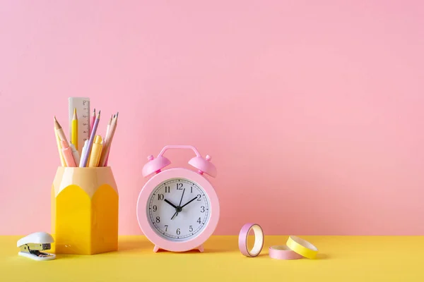 Back to school concept. Photo of school supplies on yellow desk pink alarm clock stand for pens pencils ruler stapler and adhesive tape on pink wall background