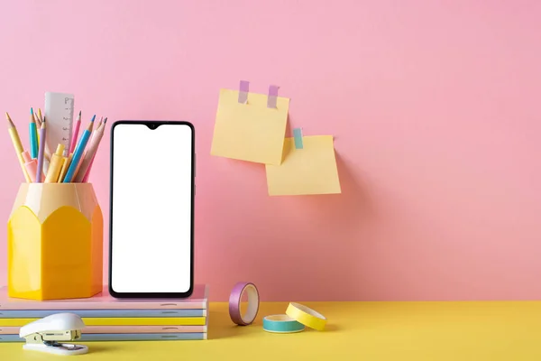 Back to school concept. Photo of school accessories on yellow desk smartphone stand for pens stapler stack of notebooks adhesive tape and sticky note paper attached to pink wall with copyspace