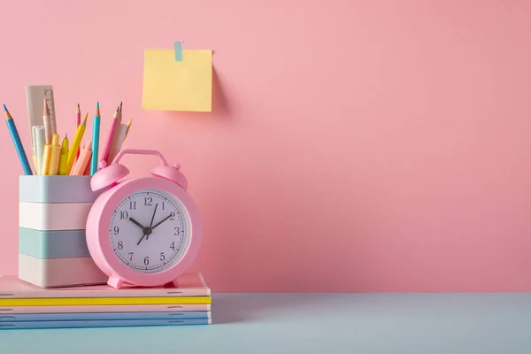 Back to school concept. Photo of school supplies on blue desk stand for pencils pens ruler alarm clock stack of copybooks and sticky note paper attached to pink wall with copyspace