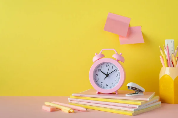 Back to school concept. Photo of school supplies on pink table alarm clock stand for pens stack of notebooks pencils mini stapler eraser and sticky notes attached to yellow wall
