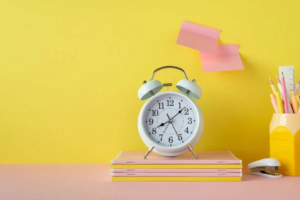 Back to school concept. Photo of school accessories on pink desk alarm clock stack of copybooks stand for pens mini stapler and sticky note paper attached to yellow wall