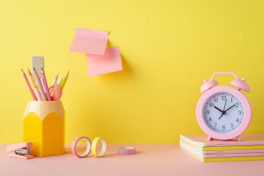 School accessories concept. Photo of alarm clock over copybooks pencil holder adhesive tape mini stapler and sticky note paper attached to yellow wall