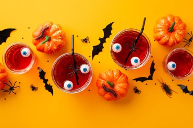 Halloween party concept. Top view photo of glasses with drink floating eyeball punch pumpkins bat silhouettes insects centipedes cockroach spiders and confetti on isolated yellow background
