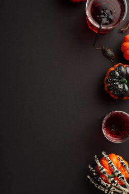 Halloween concept. Top view vertical photo of drink in glasses with floating spiders and straw skeleton hand holding pumpkin centipede and cockroach on isolated black background with copyspace