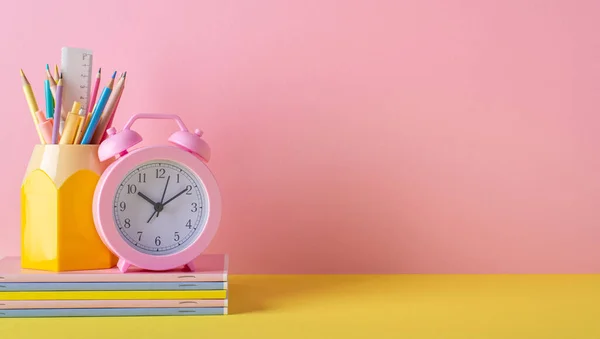 Back to school concept. Photo of school supplies on yellow desk alarm clock stand for pens pencils ruler and stack of notebooks on pink wall background with empty space