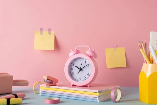 School accessories concept. Photo of color stationery on blue desk alarm clock stand for pens stack of copybooks mini stapler adhesive tape pencil-case markers sticky note paper attached to pink wall