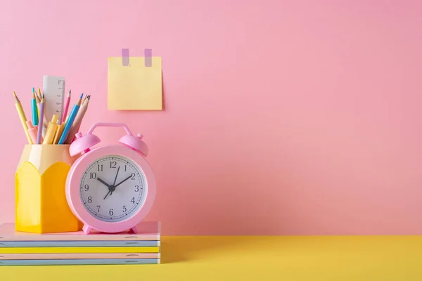 Back to school concept. Photo of school accessories on yellow desk stand for pens alarm clock stack of colorful notebooks and sticky note paper attached to pink wall with empty space