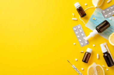 Mevsimsel hastalıklar konsepti. İlaç şişelerinin, şişelerin, termometrenin yüz maskelerinin ve kopyalanmış sarı arka planda limon kesiminin üst görüntüsü.