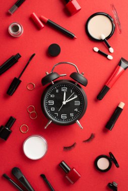 Kara Cuma konsepti. Alarm saatinin dikey fotoğrafı toz tırnak cilası göz makyajı sahte kirpikler ruj sürme dudak silindiri altın halkalar izole edilmiş kırmızı arka plan