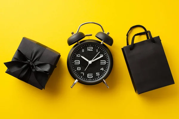 Black friday sales concept. Top view photo of black alarm clock paper bag and giftbox with ribbon bow on isolated yellow background