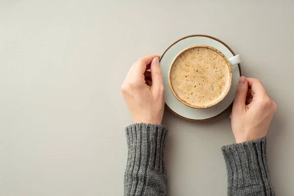 Autumn concept. First person top view photo of young woman\'s hands in knitted jumper holding saucer with cup of frothy hot drinking on isolated grey background with copyspace