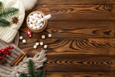 Noel arifesi kavramı. Şapka örülmüş eşarp çam dalları fincanın üst görüntüsü. Üzerinde marşmelov olan kakao fincanı, rattan plasemat viburyum üzümleri ve ahşap masa üzerinde tarçın çubukları.