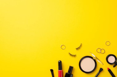 Kara Cuma konsepti. Dekoratif kozmetik fotoğrafının üst görüntüsü pudra pudrası sahte kirpikler dudak kalemi parlatıcıları ve izole edilmiş sarı arka planda altın yüzükler.
